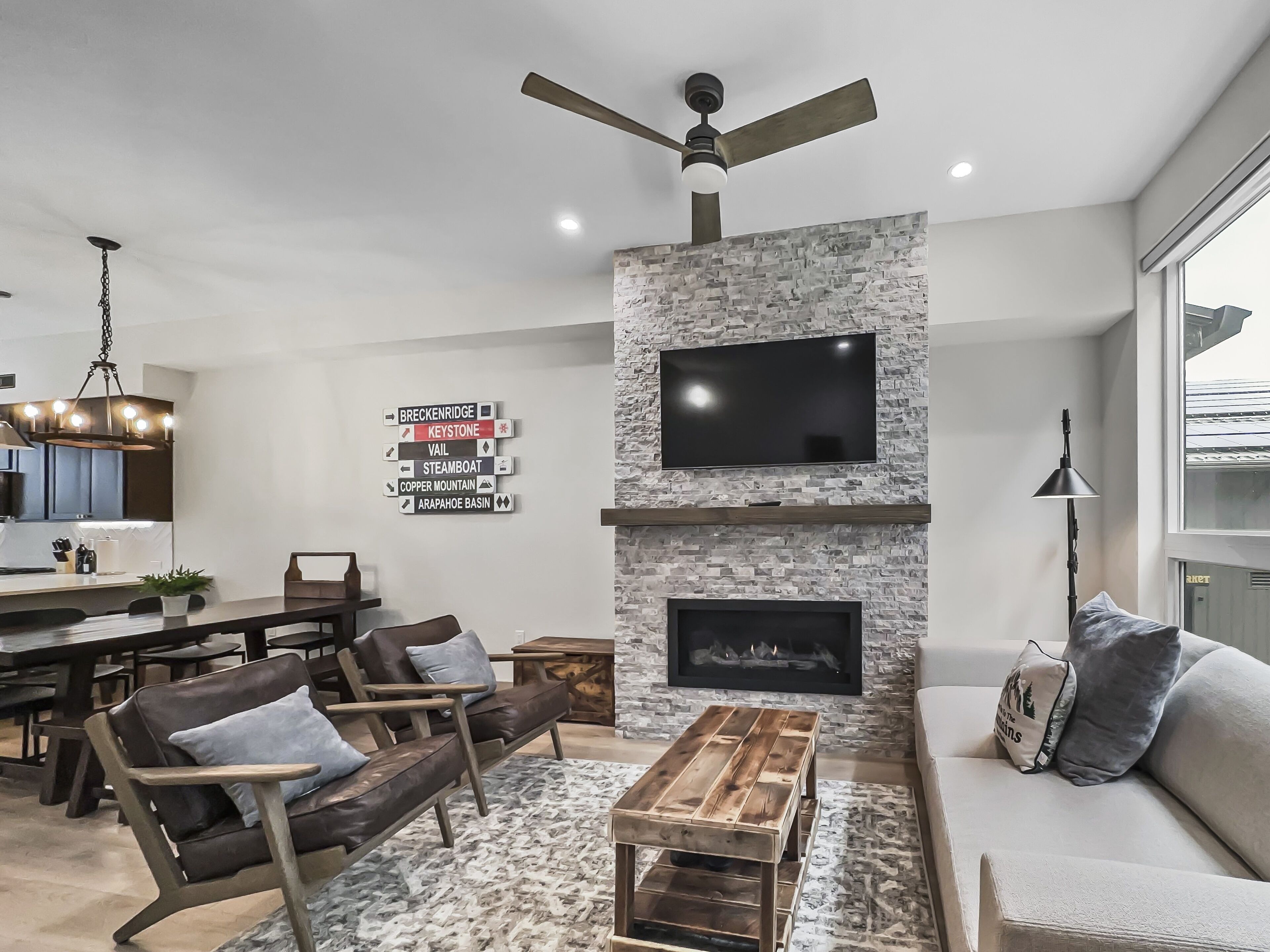 A modern living room with a stone fireplace, a flat-screen TV, and a ceiling fan.