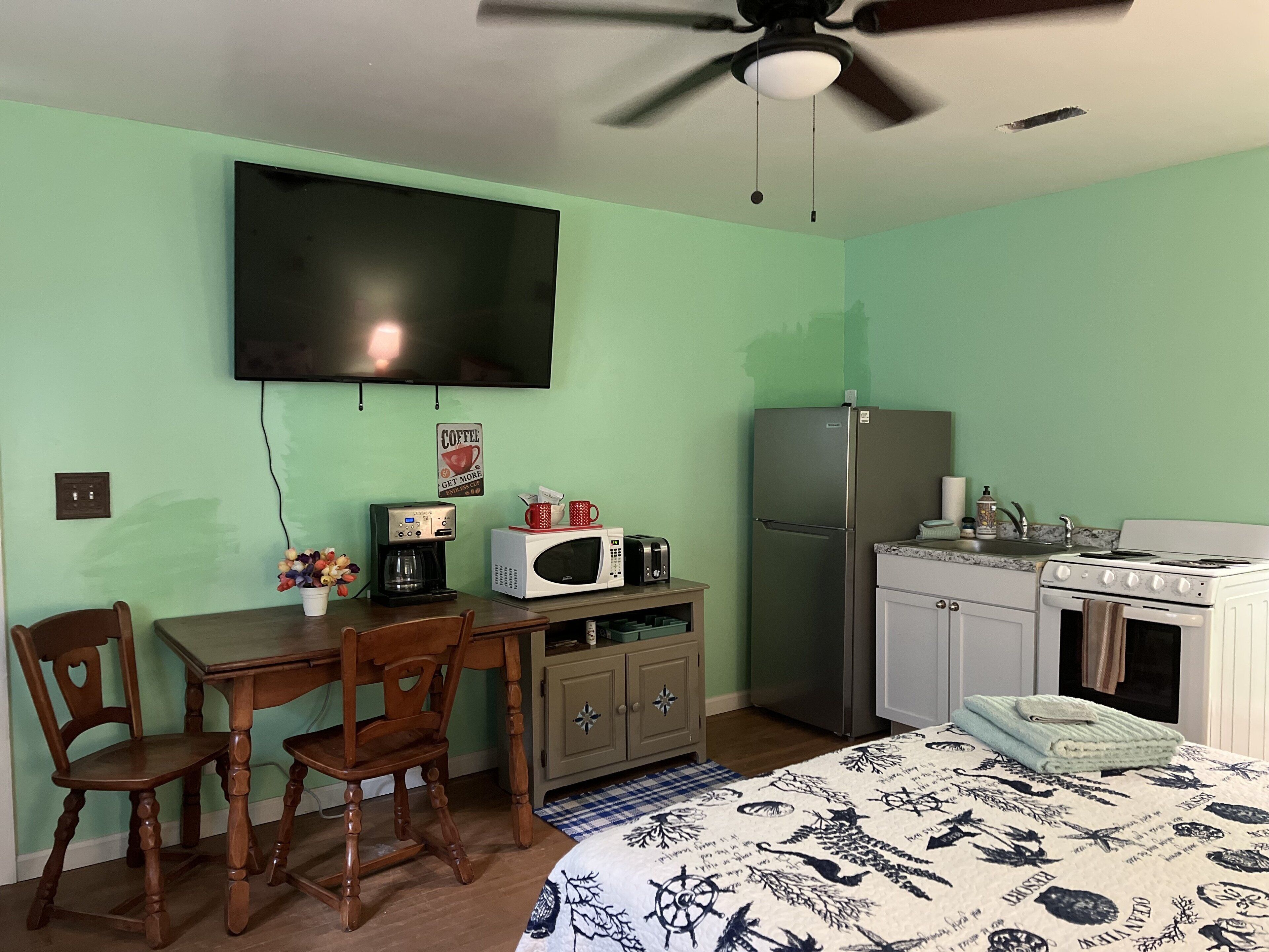 Una habitación pequeña y bien iluminada, con una pared verde, ventilador de techo, televisor de pantalla plana, refrigerador, microondas, estufa, pileta y comedor con mesa y sillas.