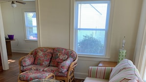 Ein Wohnzimmer mit einem Rattan-Sofa, einem gestreiften Sofa, einem Glastisch und einem Fenster mit Jalousien.