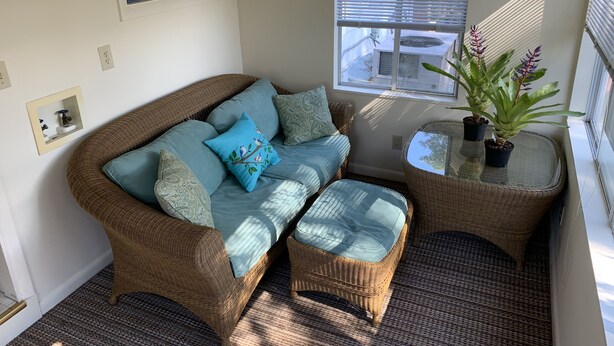 Ein gemütliches Zimmer mit einem Korbsessel und Hocker, einem kleinen Tisch mit Topfpflanzen und einem Fenster mit Jalousien.