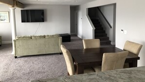 Uma sala de estar com sofá, uma mesa de jantar com cadeiras, uma TV fixada na parede e uma escada que dá acesso a outro nível.