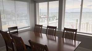 Uma sala de jantar com mesa e cadeiras de madeira, janelas amplas com persianas e vista para a água e edifícios ao longe.