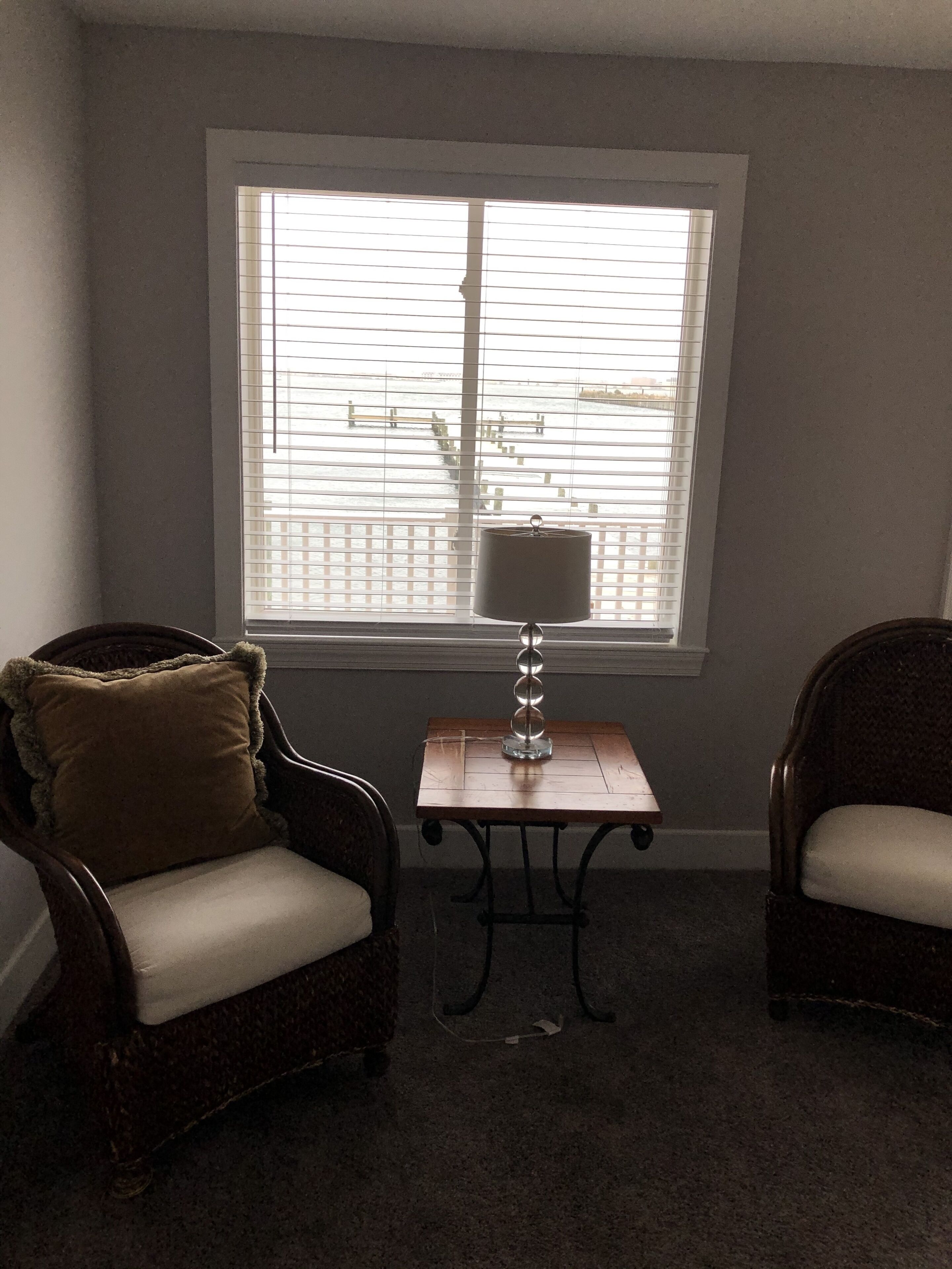 A room with two wicker chairs, a small table with a lamp, and a window with blinds.