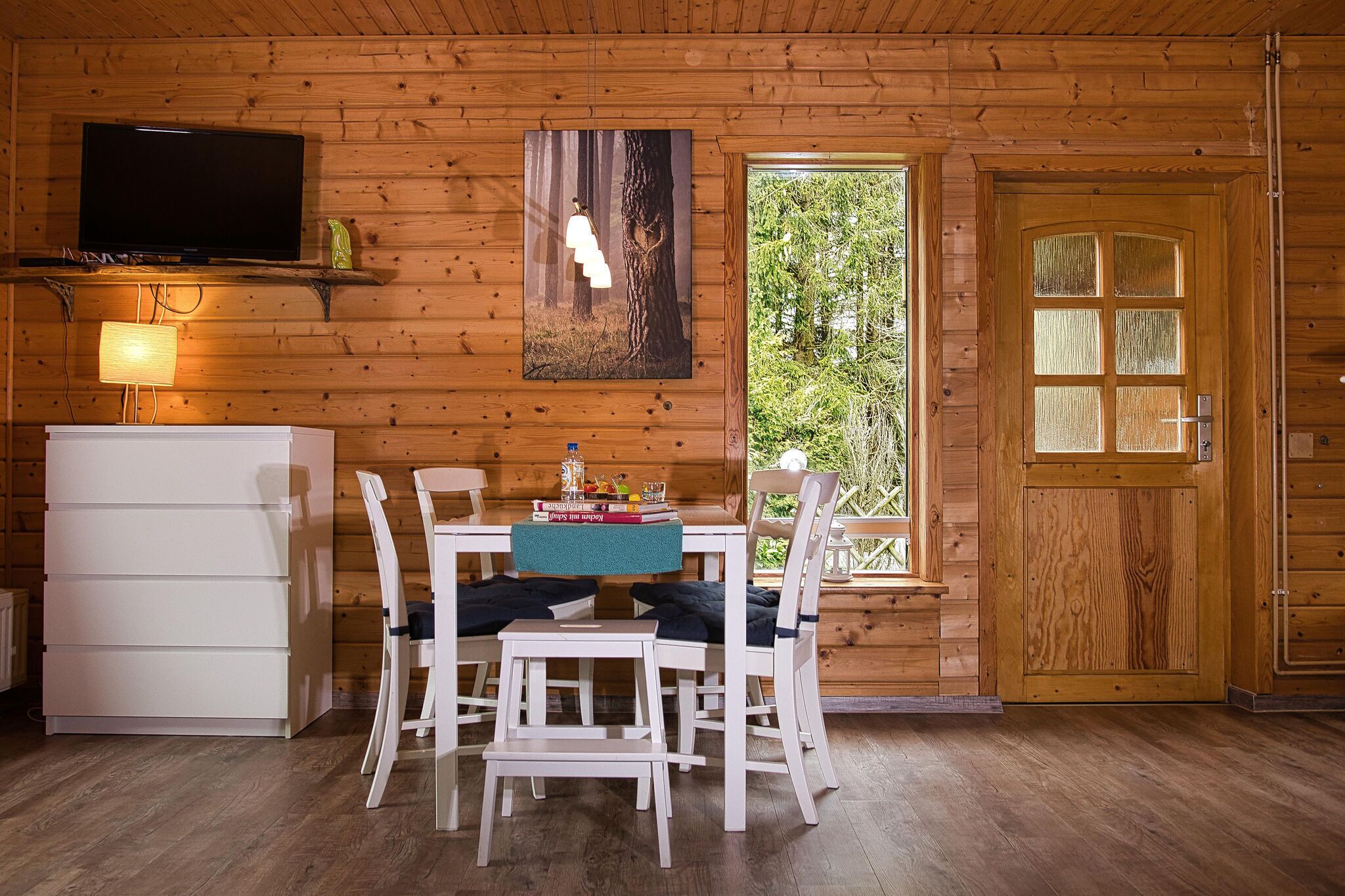 A wooden cabin interior with a dining table, chairs, a television, and a door leading outside.