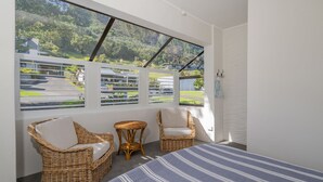 Habitación con una cama grande, dos sillas de mimbre, una mesita y vistas a un paisaje verde a través de una ventana con persianas.