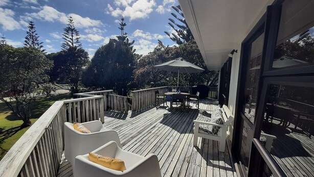 Una terraza de madera con mobiliario exterior, incluyendo sillas y una mesa, da vistas a un jardín con árboles y un cielo despejado.