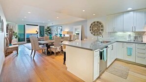 A modern kitchen with white cabinetry, a large island, and a dining area with chairs and a table. - Stunning Oceanfront Getaway at Windansea Beach (San Diego)