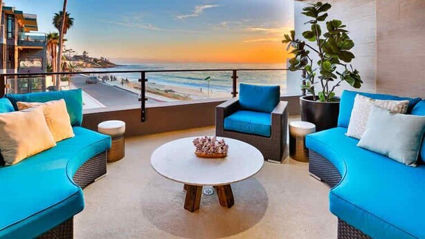 A balcony with a view of the beach and sunset, furnished with blue sofas and a round table. - Stunning Oceanfront Getaway at Windansea Beach (San Diego)