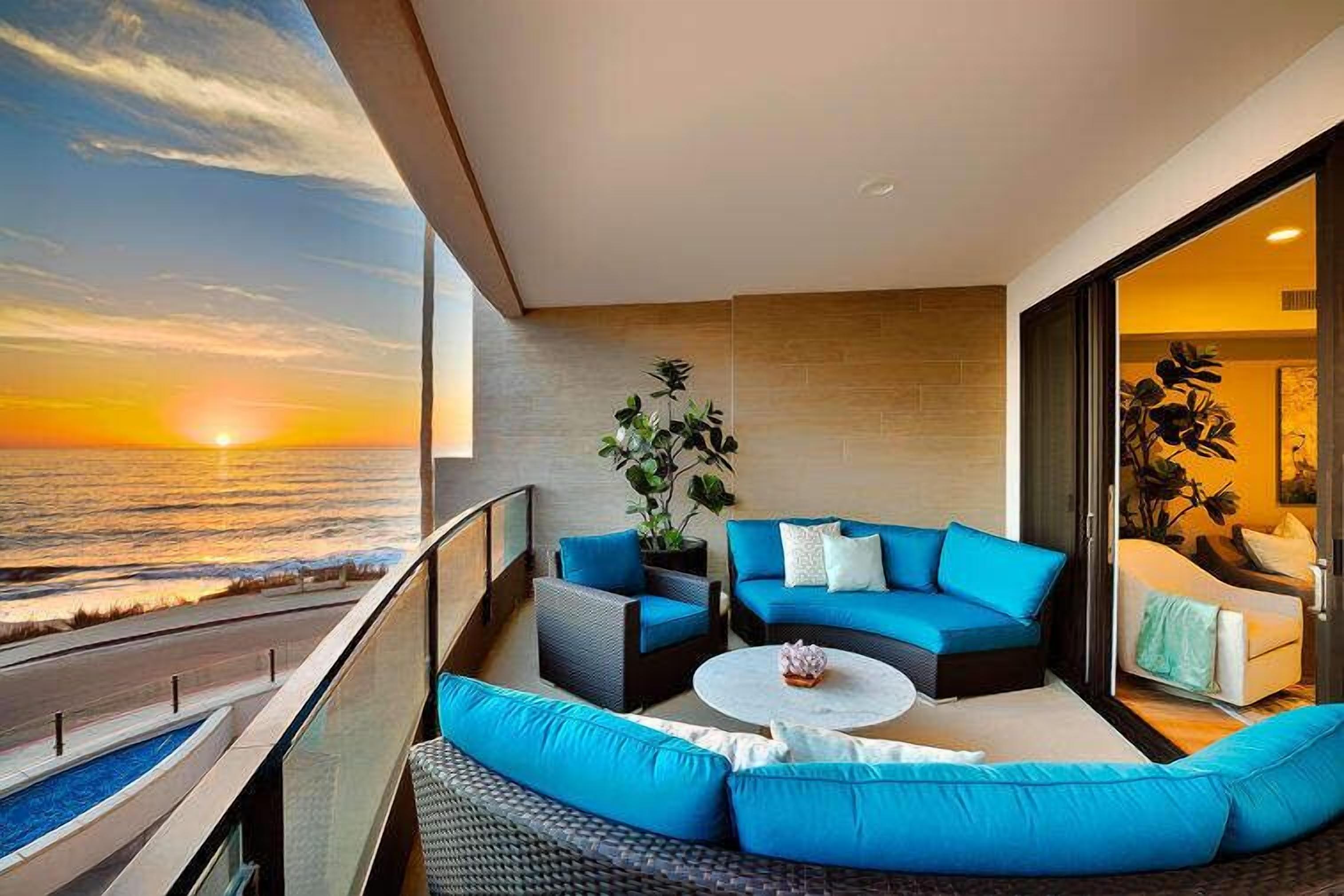Ein Balkon mit Blick auf den Sonnenuntergang über dem Meer, eine blaue Sofagarnitur, ein kleiner runder Tisch und ein Blumentopf.