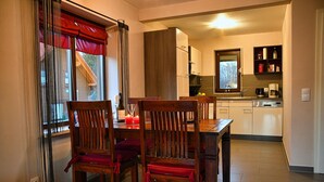 Uma área de refeições com uma mesa e cadeiras de madeira, uma janela com cortinas vermelhas e uma cozinha ao fundo.