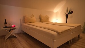 Uma cama de casal com roupa de cama bege, uma mesa de cabeceira com uma garrafa e copos, e um pequeno vaso com flores.