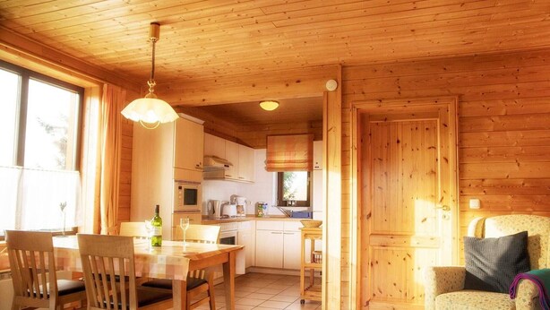 Interior em madeira com mesa de jantar, cadeiras, espaço de cozinha e área de estar aconchegante.