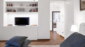 A modern living room with a flat-screen TV, built-in shelving with books and decorative items, a sofa with cushions, and a kitchen area in the background. - Country House "IL CERRO" (Toscana)