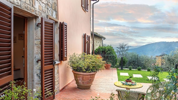 A stone building with wooden shutters, a potted plant, and a table with chairs on a paved patio. - Country House "IL CERRO" (Toscana)