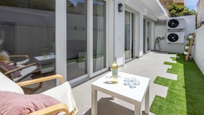 Une terrasse extérieure moderne, avec une porte vitrée donnant sur un balcon, une petite table avec une bouteille et des verres, et un espace recouvert d’herbe artificielle verte.