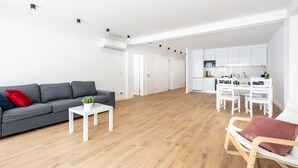 Un vaste espace de vie comprenant un canapé gris, une table basse blanche et un plancher en bois. On y trouve également une cuisine avec des meubles blancs et une table à manger.