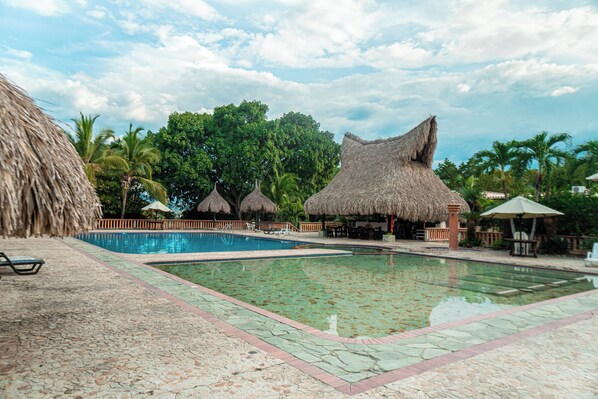 Un área de piscina con estructuras de techo de paja, palmeras y un cielo azul despejado.