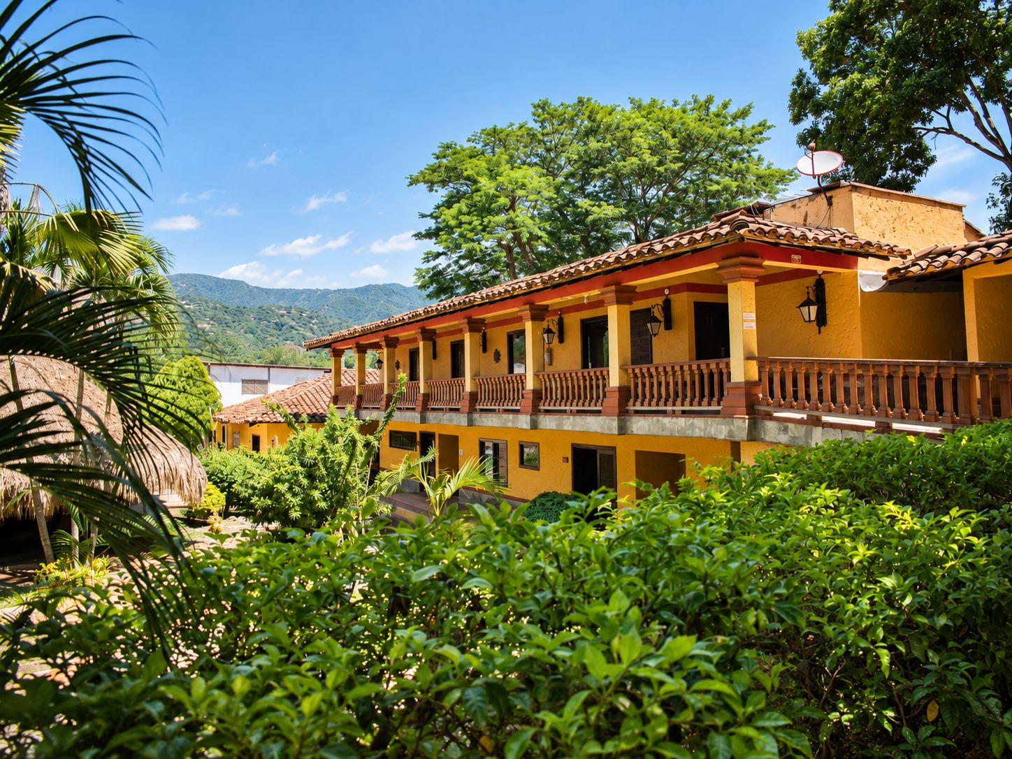 Um edifício de dois andares com varandas, rodeado por vegetação exuberante e um céu claro.
