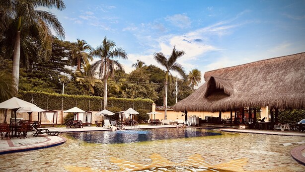 A pool area with lounge chairs and umbrellas, surrounded by palm trees and a thatched-roof building. - Hotel Porton del Sol  (Santa Fe de Antioquia)