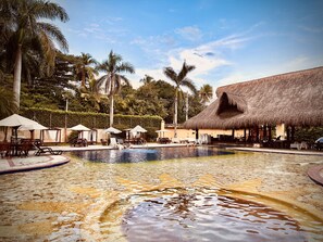 A pool area with lounge chairs and umbrellas, surrounded by palm trees and a thatched-roof building. - Hotel Porton del Sol  (Santa Fe de Antioquia)