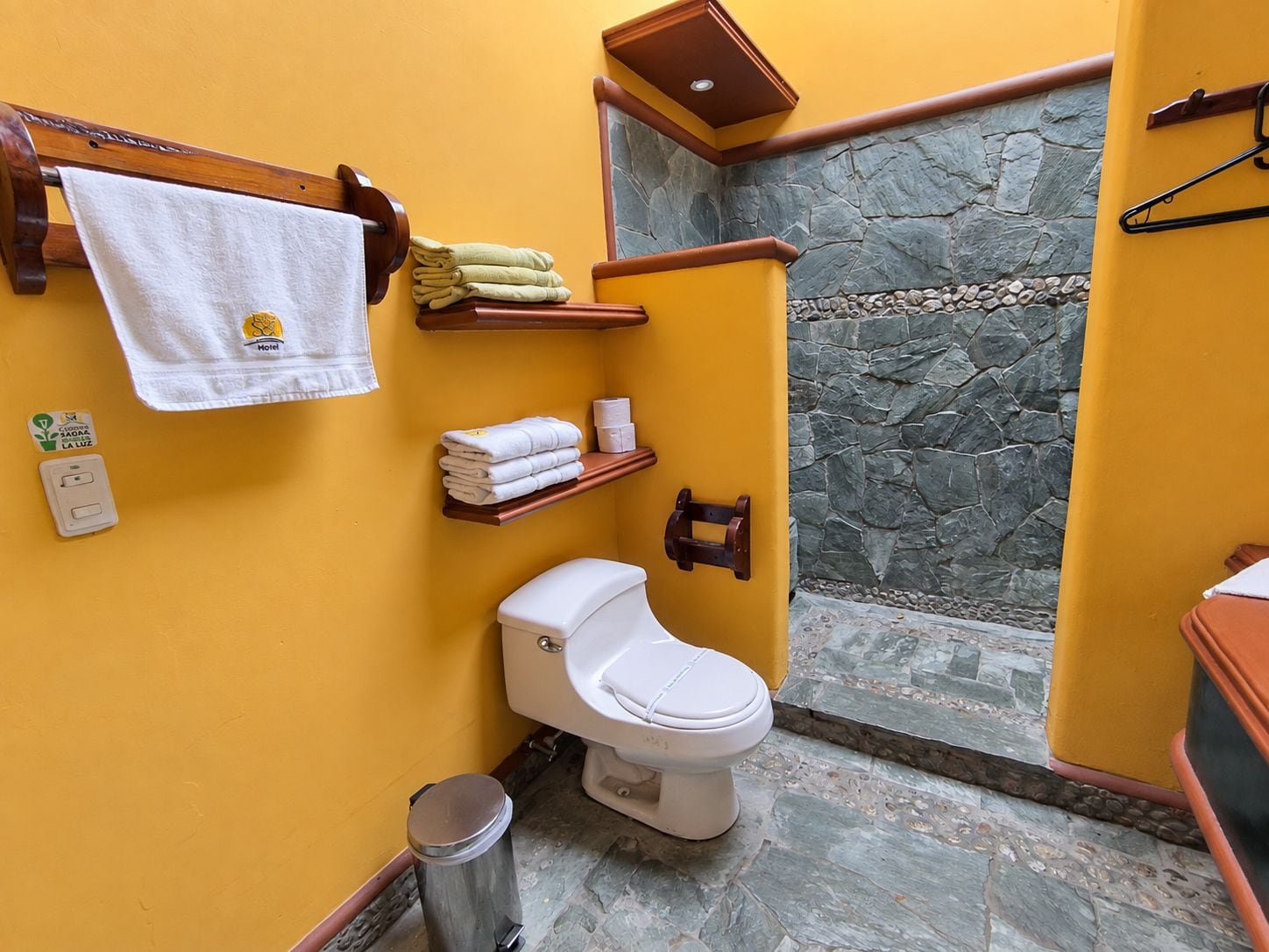 Une salle de bain comprenant des toilettes, un lavabo, un porte-serviettes avec des serviettes, une poubelle et un espace douche avec des murs en pierre.