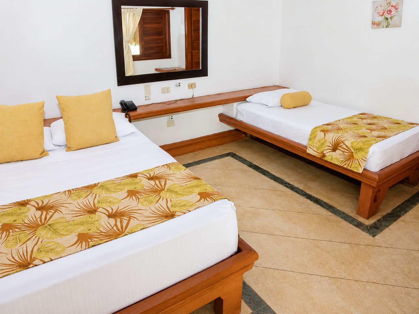 Quarto de hotel com duas camas de solteiro, um banco de madeira, uma mesinha, um espelho e um edredom com padrão floral.
