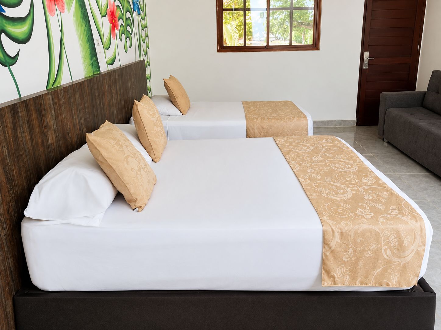 A hotel room with two beds, a sofa, a window with a view of greenery, and a wall with a floral mural.
