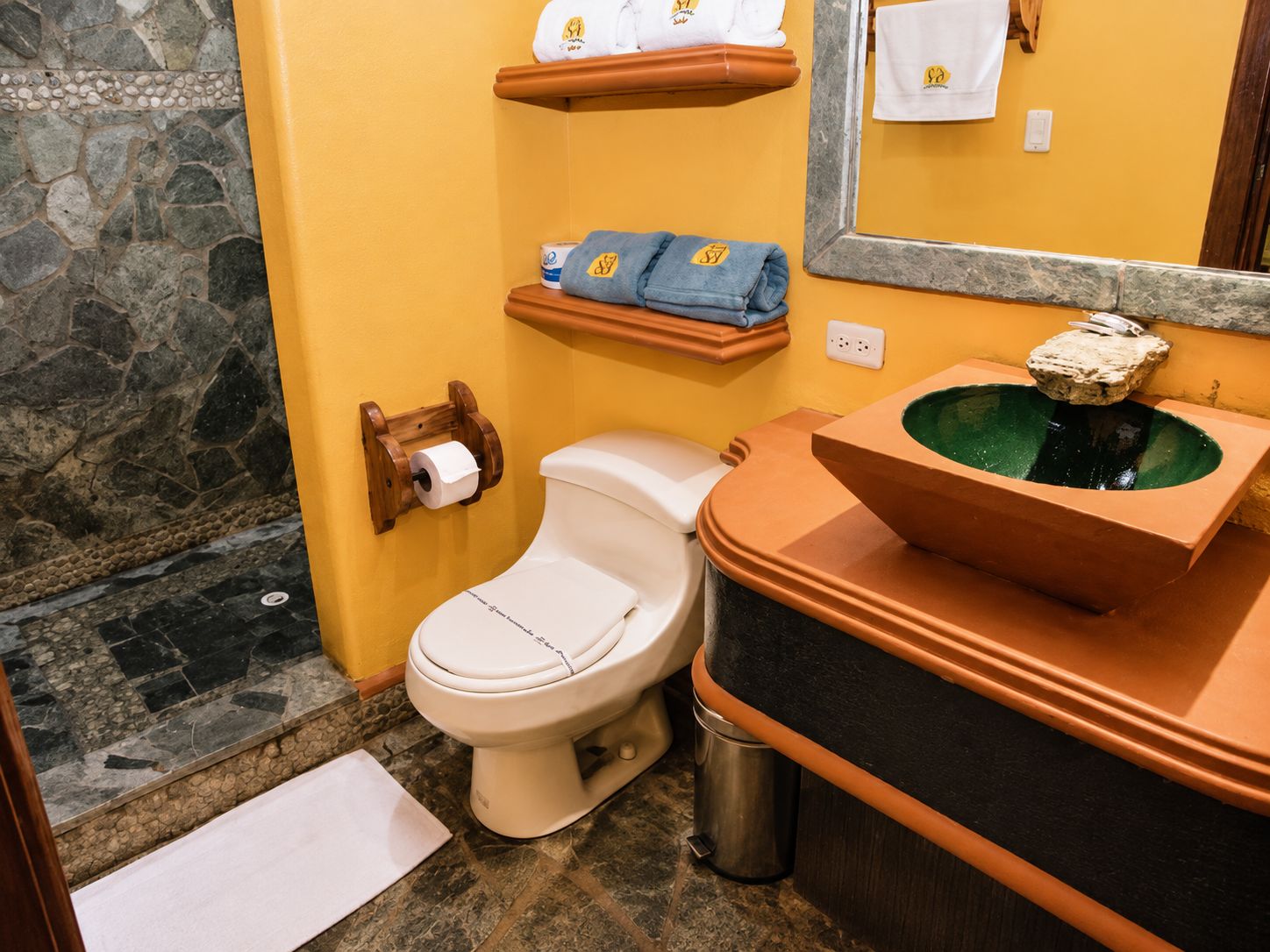 A bathroom with a toilet, a sink with a stone basin, a towel rack with towels, and a mirror.