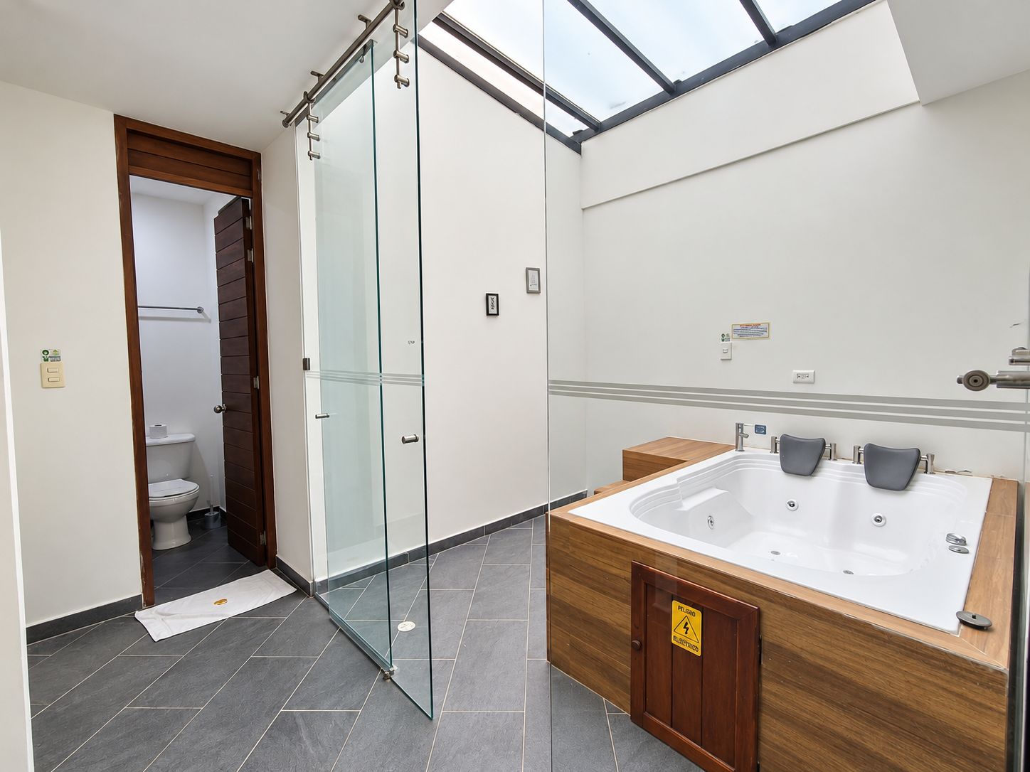 Une salle de bain moderne avec une douche à l’italienne en verre, un meuble-lavabo en bois avec une baignoire, et une lucarne au-dessus.