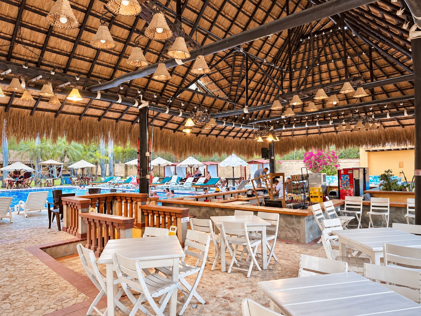 An outdoor pool area with lounge chairs and tables, a bar, and a thatched roof structure.
