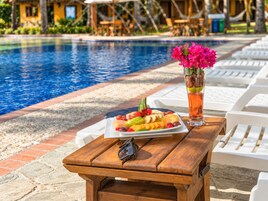 Una mesa de madera con una bandeja de frutas y una bebida, junto a un área de descanso junto a la piscina.