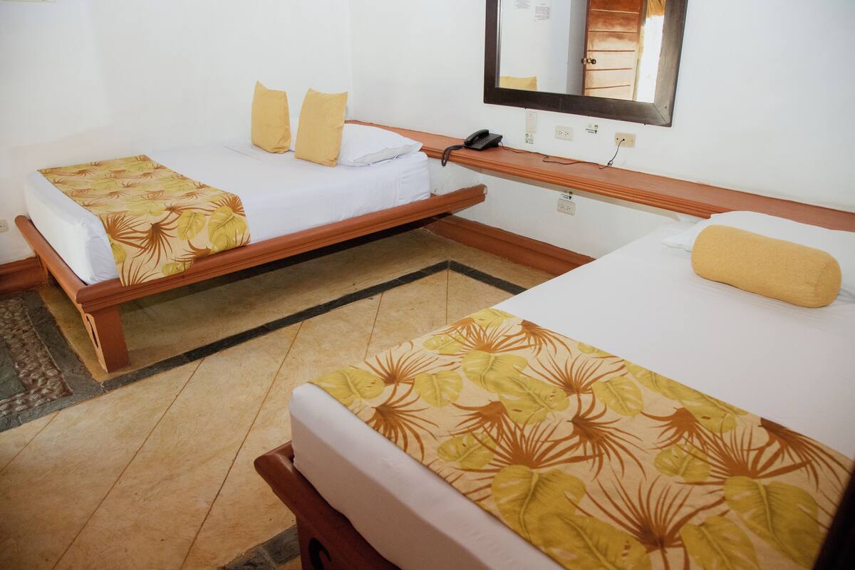 A hotel room with two single beds, each with a floral-patterned bedspread and a wooden headboard. There is a wooden nightstand between the beds, and a mirror above the headboard on the right side.