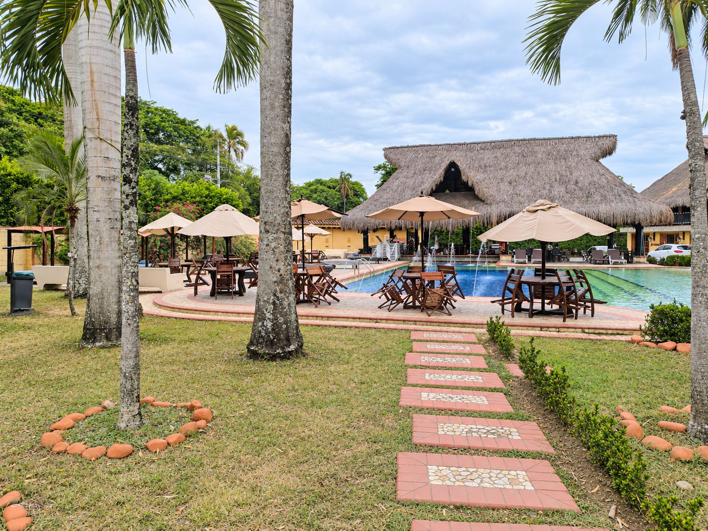 A pool area with thatched-roof structures, lounge chairs, and a paved walkway.