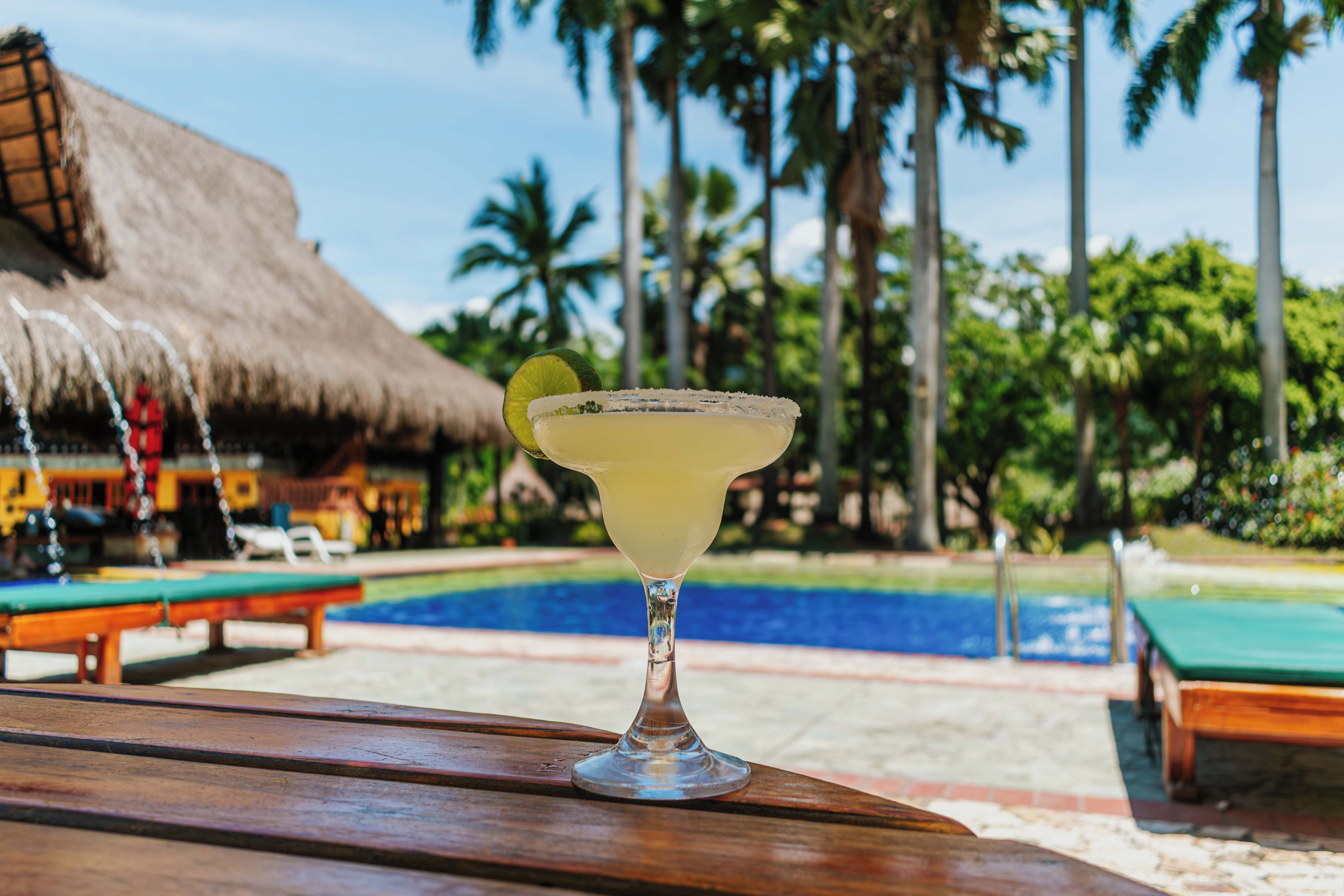 A cocktail on a wooden table with a pool and palm trees in the background.