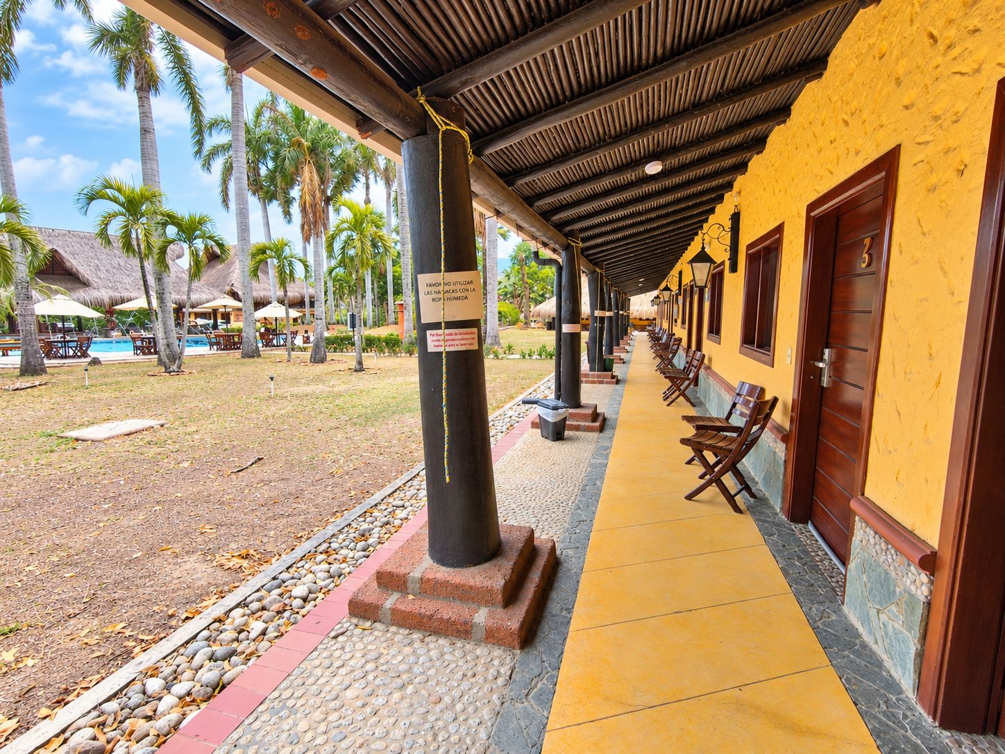 A covered outdoor seating area with a pool in the background, wooden benches, and a yellow building facade.