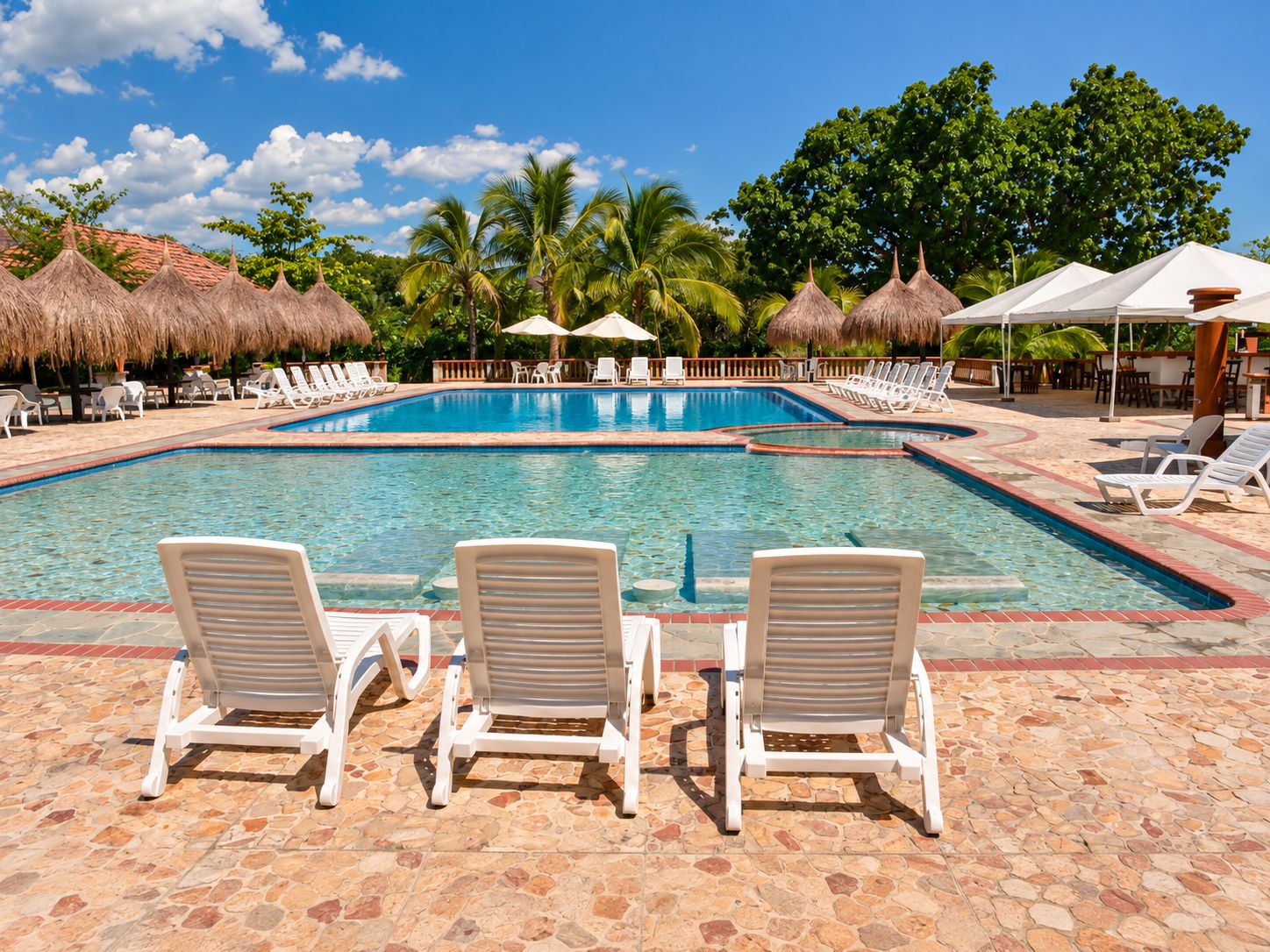 Una piscina con sillones de descanso y sombrillas, rodeada de palmeras y sombrillas de paja.