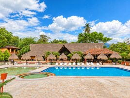 Una piscina con sillas de descanso y sombrillas de paja, rodeada de vegetación tropical y edificios con techos de tejas.