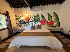 Un dormitorio con una cama grande, una ventana con cortinas, un mural en la pared con un loro y hojas tropicales, y un banco con cojín.