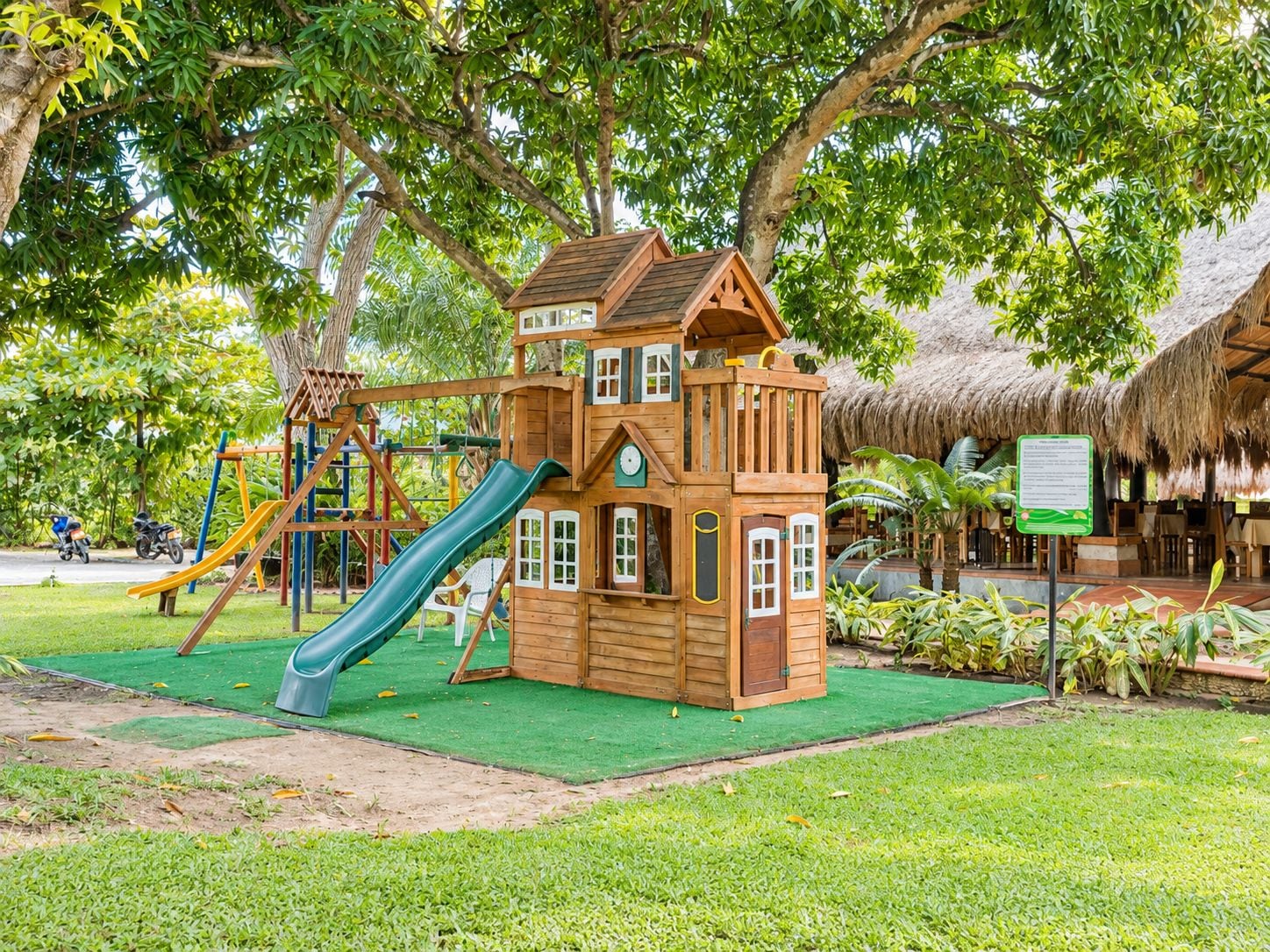 A wooden playhouse with a slide, surrounded by lush greenery and a thatched-roof structure.