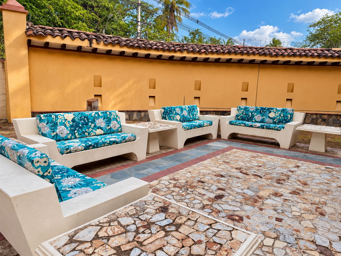 An outdoor seating area with white sofas and blue floral cushions, a stone-tiled floor, and a yellow wall with a tiled roof.