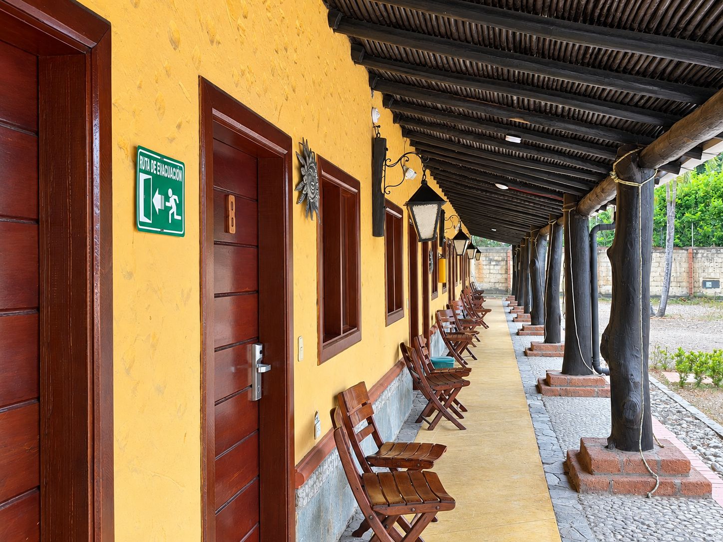 A corridor with wooden doors and benches, a green evacuation route sign, and a covered area with columns.