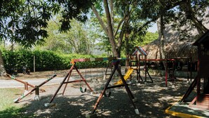 A playground with swings and a slide, surrounded by trees and a thatched-roof structure. - Hotel Porton del Sol  (Santa Fe de Antioquia)