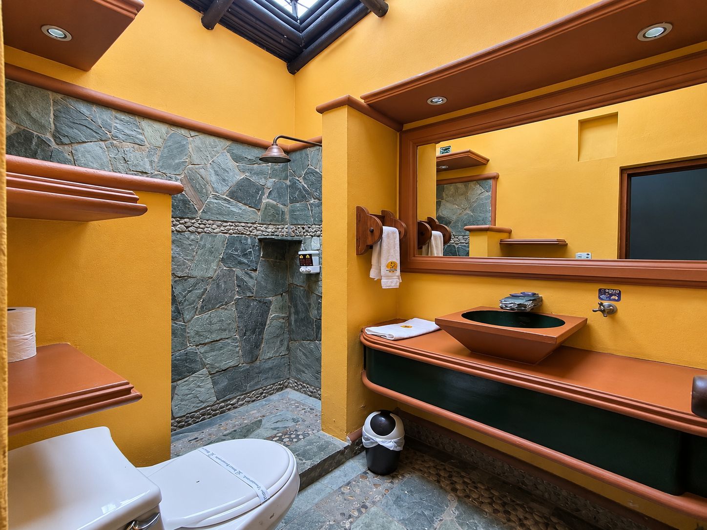 Ein Badezimmer mit Duschkabine aus Stein, Toilette, Waschbecken und einem Spiegel mit Holzrahmen.