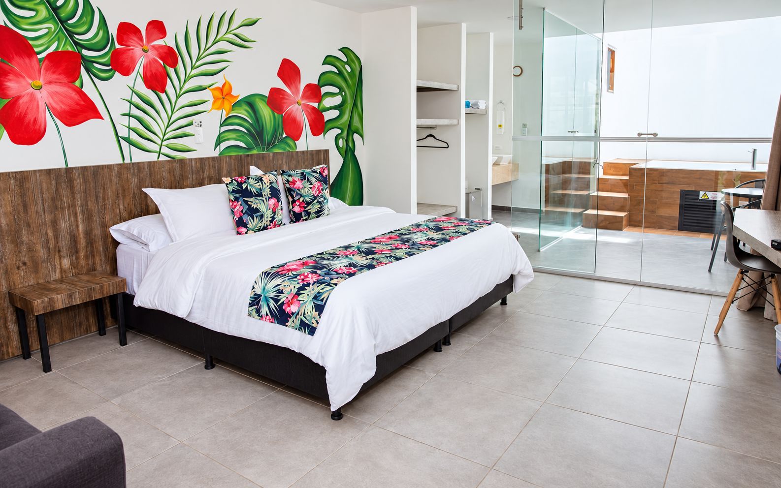 Habitación de hotel moderna con una cama, un pequeño banco de madera, un mural temático tropical y un ventanal de vidrio que da a un balcón.