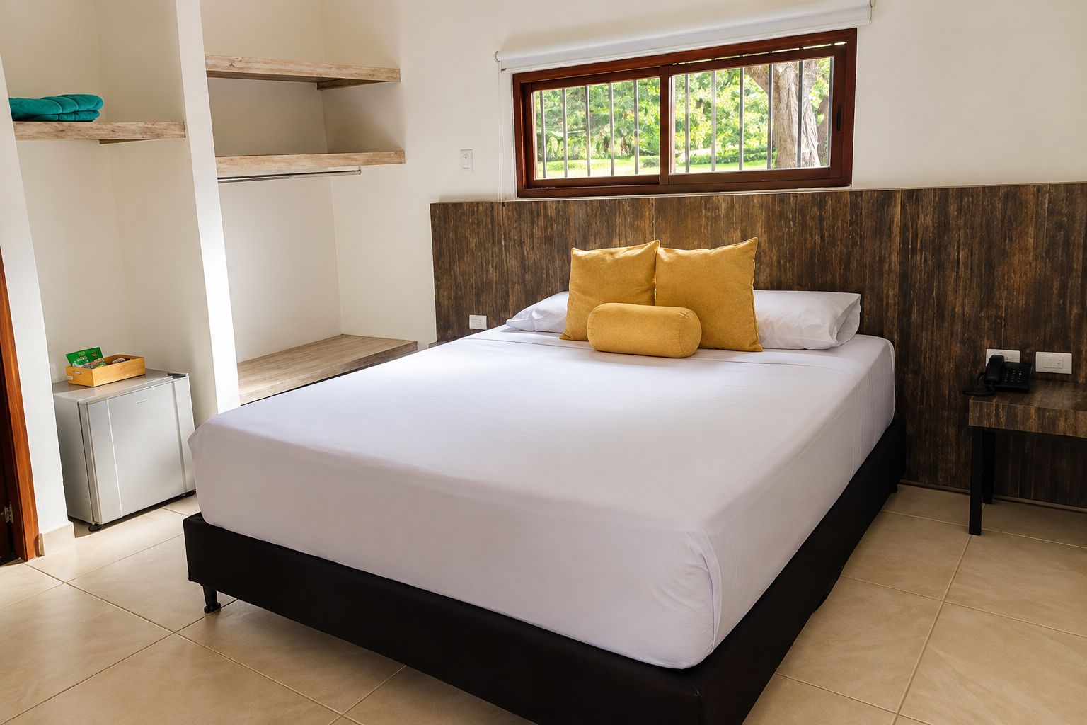 Quarto de hotel com cama, escrivaninha, mini-geladeira e janela com vista para área verde.