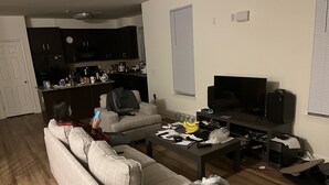 A living room with a sofa, coffee table, TV, and kitchen area in the background.