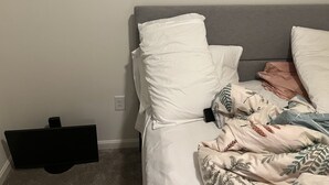 A bedroom with a bed, pillows, a blanket, a TV, and a power outlet on the wall.