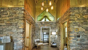 A stone-walled hallway with a wooden ceiling, a chandelier, a chair, and a bed. - 4 BR w Views, Private trails, hot tub, 3 king beds, EV Charger (Mineral Bluff)