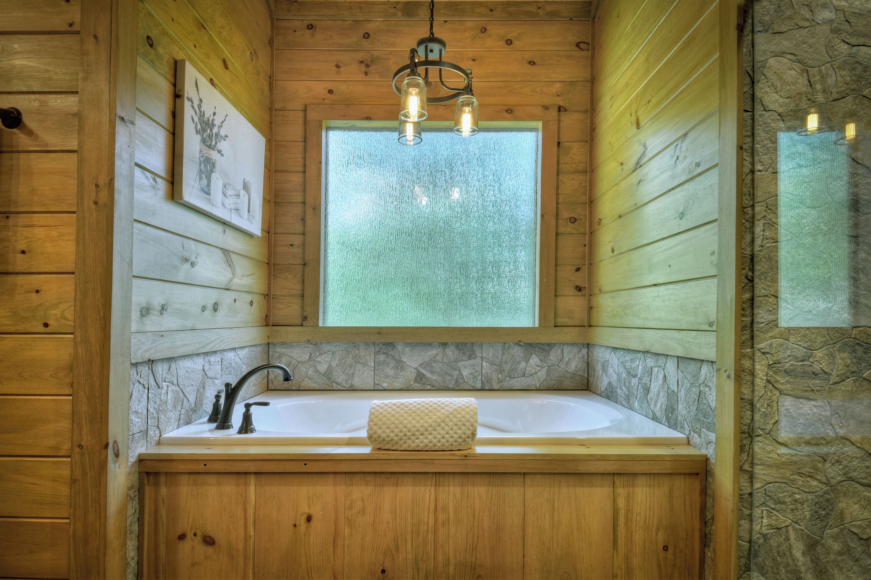 Une salle de bain rustique, située dans une cabane en bois, avec un mur en pierre, une baignoire en bois, un luminaire suspendu et un tableau encadré accroché au mur.