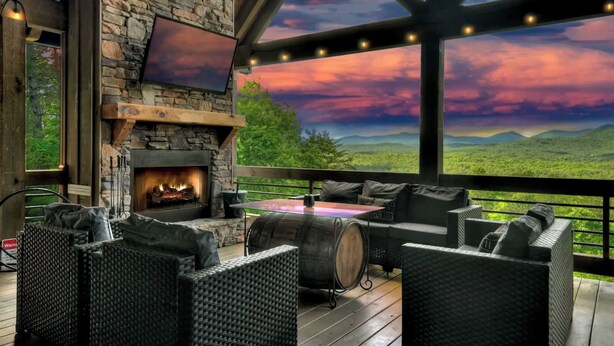A cozy outdoor seating area with wicker furniture, a stone fireplace, and a large window offering a scenic view of the mountains. - 4 BR w Views, Private trails, hot tub, 3 king beds, EV Charger (Mineral Bluff)